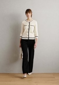 Cardigan blanc avec des bordures et des boutons noirs, associé à un pantalon noir. Modèle tenant un sac blanc, se tenant sur un sol en bois clair.