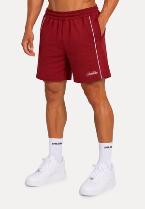 Uomo che indossa pantaloncini sportivi color granata con strisce laterali bianche e scarpe da ginnastica bianche con calzini firmati, in piedi con le mani rilassate.