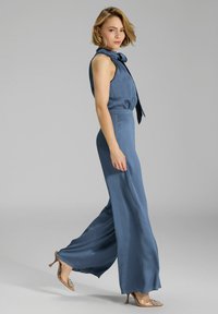 Blauwe jumpsuit met een aansluitend lijfje en brede, vloeiende broekspijpen. Heeft een schouderstrikdetail en een textuurstof. Compleet met hoge hakken.