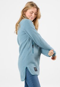 Pullover in pile azzurro chiaro con un'esterno texturizzato e morbido, tasca con zip, colletto alto e spacchi laterali per facilitare il movimento.