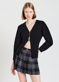 Cardigan nero lavorato a maglia con scollatura a V profonda e zip frontale, abbinato a una gonna plissettata a quadri grigia. Texture liscia, vestibilità aderente e design moderno.