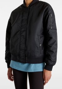 Blouson aviateur noir avec une texture lisse, des poignets et une taille côtelés. Comprend une poche zippée sur la manche gauche et des poches latérales.