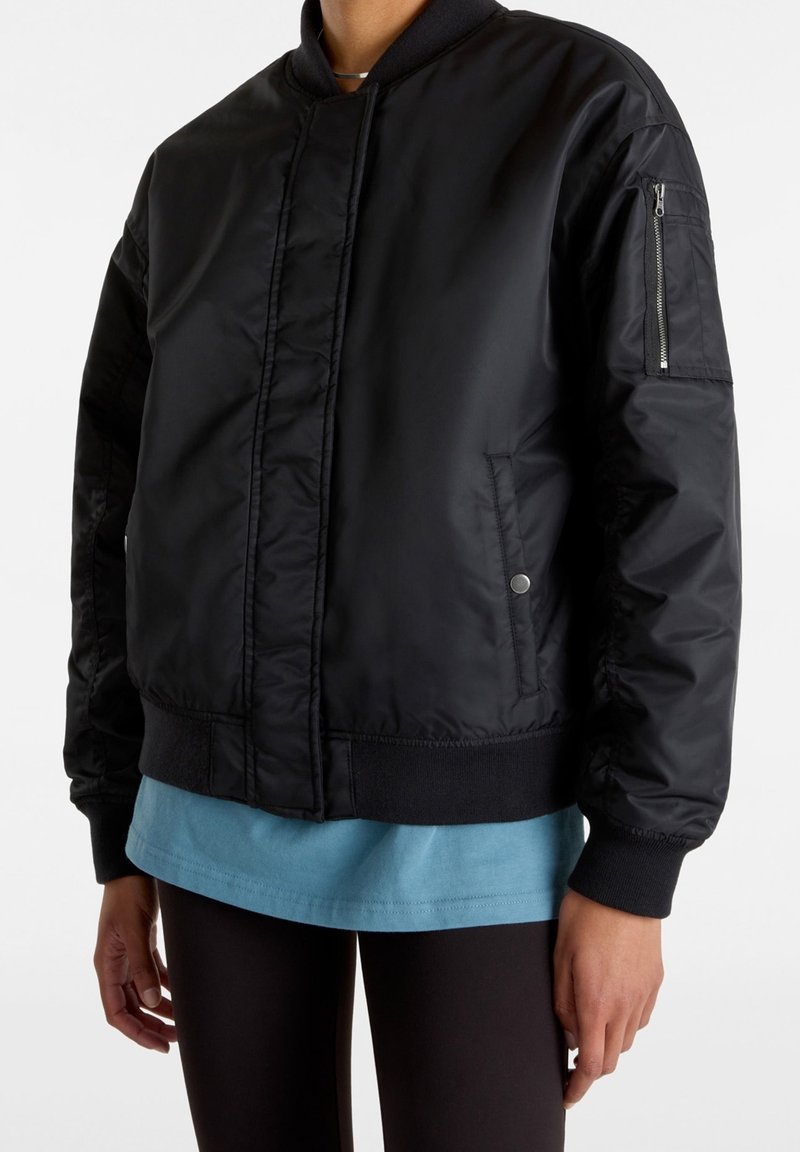 Blouson aviateur noir avec une texture lisse, des poignets et une taille côtelés. Comprend une poche zippée sur la manche gauche et des poches latérales.