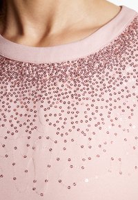 Lyserød skjorte med rund halsudskæring, der har en tekstureret overflade prydet med skinnende perler i et spredt mønster og en let glans.