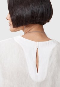 Hvid bluse med rund hals, nøglehul-detalje bagpå og et let tekstureret stof. Højden er glat og delikat.