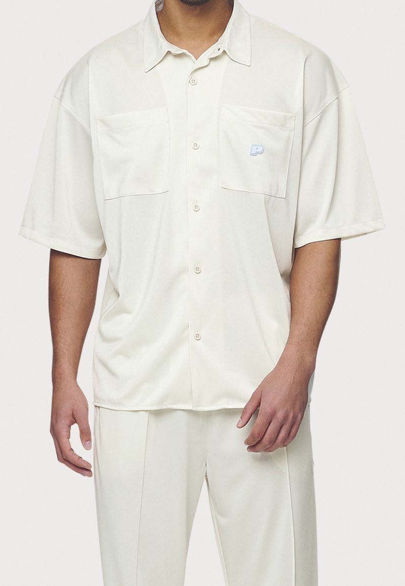 Biała koszula z krótkim rękawem wykonana z gładkiej tkaniny. Posiada dwie przednie kieszenie oraz małe logo. Zapięcie na guziki z przodu.