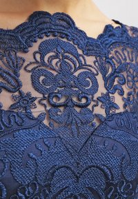 Modré květinové krajkové vyšívání na průhledné látce přes hruď, zobrazující složité vířící a květinové vzory v detailním záběru.