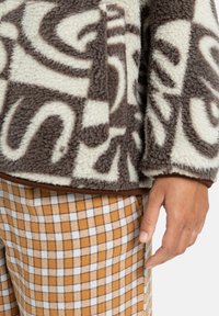 Veste en polaire floue crème et marron à motifs avec un ourlet marron, associée à un pantalon à carreaux orange et blanc. Main posée sur le côté.