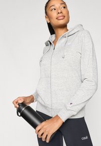 Γυναίκα που φοράει γκρι φούτερ με φερμουάρ και μαύρο κολάν, κρατώντας μαύρο παγούρι, κοιτάζοντας προς τα πάνω και πλάγια.