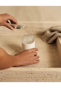 Mani che versano un liquido da una piccola bottiglia trasparente in una bottiglia più grande con superficie satinata, su una superficie di pietra beige con un tessuto testurizzato nelle vicinanze.