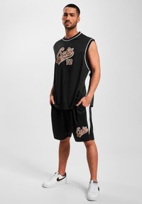 T-shirt de sport sans manches noir avec des accents blancs, arborant un logo graphique marron. Associé à un short noir et des baskets blanches avec des détails noirs.