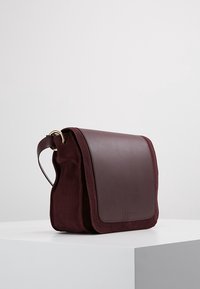 Borsa in pelle bordeaux con finitura scamosciata. Forma rettangolare, pattina ripiegabile e tracolla regolabile. Accenti in metallo dorato visibili.