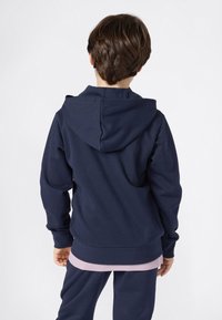 Niño con cabello corto castaño, vistiendo una sudadera azul marino y pantalones a juego, de pie de espaldas contra un fondo blanco liso.