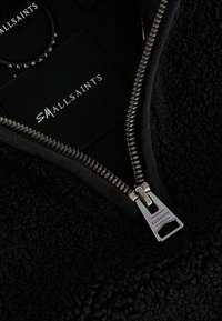 Juodas flisinis zip-up džemperis su sidabriniu užtrauktuku ir prekės ženklo žyme, turintis tekstūruotą paviršių ir kontrastingą medžiagą apykaklės viduje.