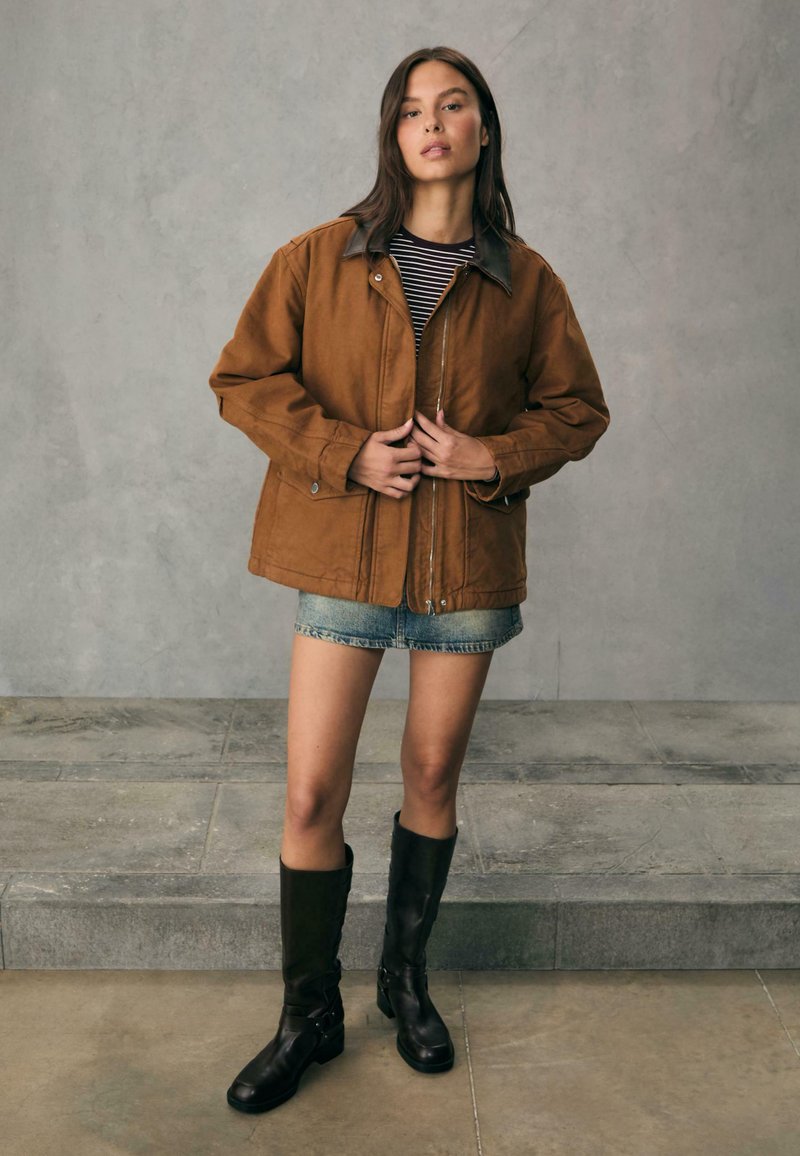 Brun, oversized jakke med en kontrasterende krave, parret med en stribet skjorte, denim shorts og knæhøje sorte støvler. Glat tekstur.