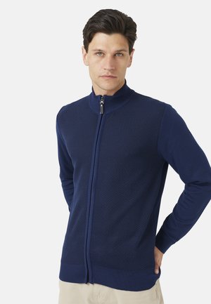 Jonge man met donker haar, met een marineblauwe trui met ritssluiting, met een gestructureerde voorkant en effen mouwen, staand tegen een effen achtergrond.