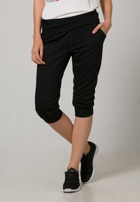 Pantalones deportivos negros recortados con una cinturilla ancha, bolsillos laterales y puños acanalados, combinados con zapatillas negras sobre un fondo neutro.