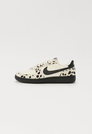 Sneaker bianca con motivo a macchie nere, logo Nike Swoosh nero, lacci piatti e suola in gomma nera. Design classico a basso profilo.