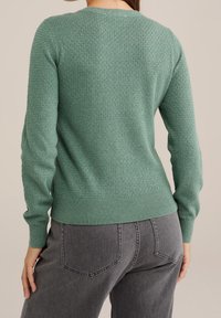 Woman wearing a textured green long-sleeve sweater and gray high-waisted jeans, shown from the back against a neutral background.