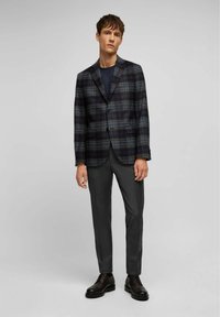 Geruite blazer in donkere tinten, met een notch lapel, twee knopen en klepzakken. Gecombineerd met slanke grijze broeken en zwarte schoenen.