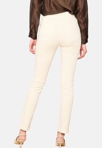 Kreminių spalvų prigludę kelnių, su lygiu tekstūru, su dviem užpakalinėmis kišenėmis ir vidutinio aukščio juosmens dizainu. Suderinta su permatomu rudos spalvos palaidiniu ir aukštakulniais.