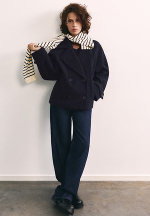 Jeune femme aux cheveux bouclés portant un manteau croisé bleu marine, un jean à jambes larges, des chaussures noires et une écharpe rayée tenant une extrémité.