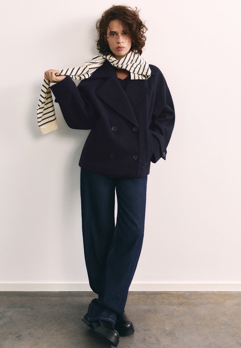 Jeune femme aux cheveux bouclés portant un manteau croisé bleu marine, un jean à jambes larges, des chaussures noires et une écharpe rayée tenant une extrémité.