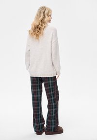 Blondynka z falującymi włosami, ubrana w luźny beżowy sweter, zielono-bordowe spodnie w kratę i brązowe wsuwane buty, stoi zwrócona tyłem.