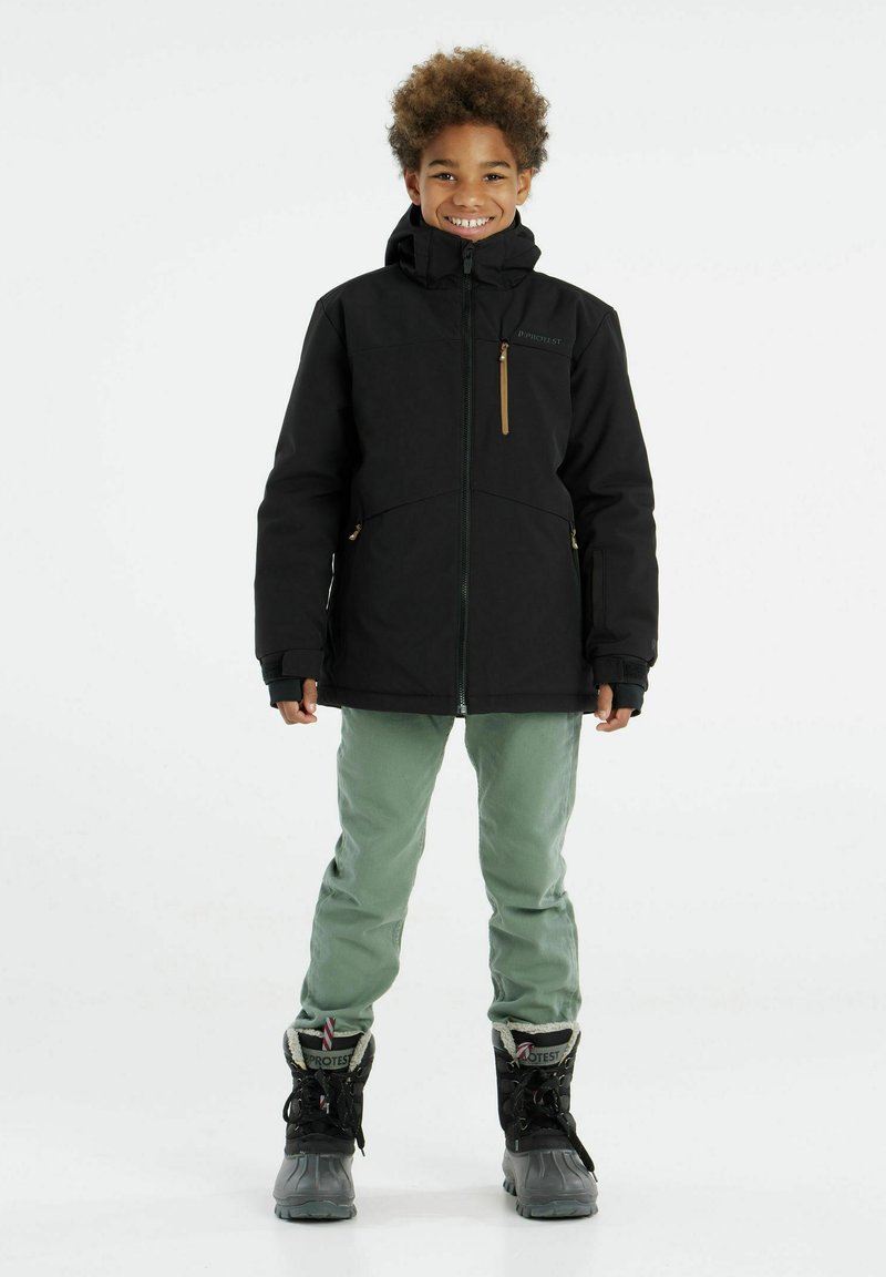 Schwarze Winterjacke mit Kapuze, Reißverschluss vorne und seitlichen Taschen, kombiniert mit grünen Hosen und schwarzen Stiefeln, mit Kunstfellfutter.
