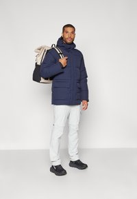 Marineblå pufferjakke med hette, flere frontlommer, sammen med lysegrå cargobukser og svarte sporty sko. Ryggsekk i beige og svart.