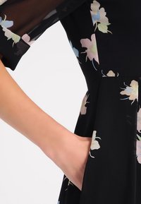 Svart blommig klänning med korta ärmar, prydd med pastellfärgade blommönster. Tyget verkar lätt och har en fickdetalj på sidan.