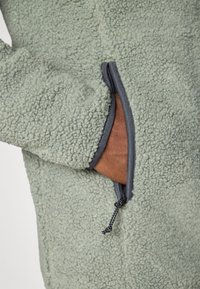 Giacca in pile verde chiaro con una superficie testurizzata, dotata di una tasca con zip con bordo grigio scuro e un accento in pelle marrone all'interno.