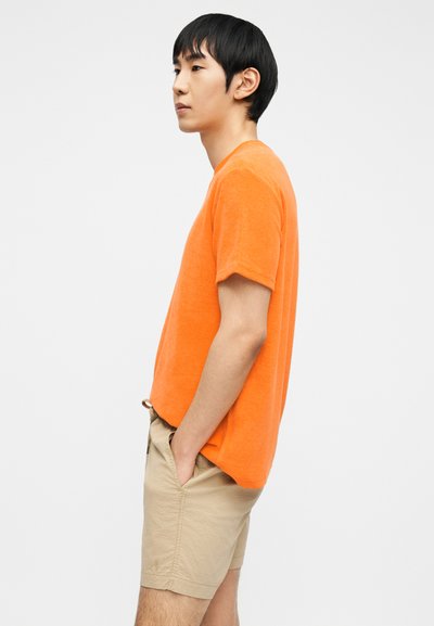 Νέος άνδρας όρθιος πλαγίως με τα χέρια στις τσέπες, φοράει πορτοκαλί κοντομάνικο πουκάμισο και μπεζ σορτς, σε απλό φόντο.