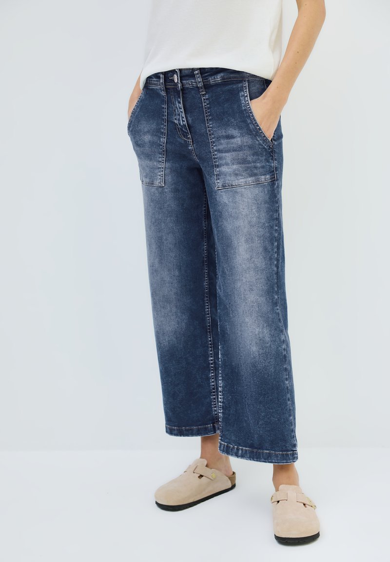 Weite Jeans in verblasst dunkelblauem Denim, mit vorderen Taschen und Knopfverschluss, kombiniert mit beigen Schlupf-Clogs.