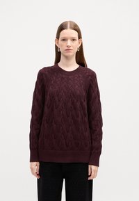 Gilės mezgimo megztinis giliu burgundišku atspalviu, su apvalia apykakle ir raukšlėmis rankogaliuose. Dėvimas su tamsiomis, tekstūruotomis kelnėmis.