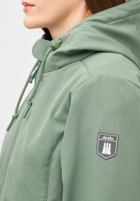 Groene hoodie met een gladde textuur, voorzien van een rits en verstelbaar trekkoord. Toont een zwart-wit logo-patch op de mouw.