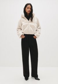 Kremna zip-up jakna iz umetnega krzna s skrajšano zasnovo, v kombinaciji s črnimi hlačami širokega kroja z vrvico in elegantnimi črnimi petami.