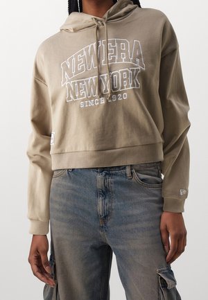 Beżowa krótka bluza z kapturem z napisem „New Era New York Since 1920” noszona z wypranymi niebieskimi dżinsami z kieszeniami typu cargo.
