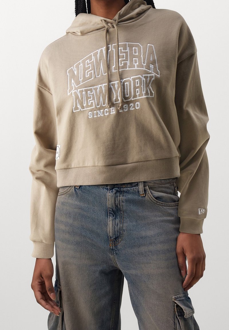 Sweat à capuche court beige avec le texte "New Era New York Since 1920", porté avec un jean bleu délavé avec poches cargo.