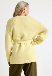 Lys gul oversized strikcardigan med bælte i taljen og ribbede manchetter. Blød tekstur, afslappet pasform og mellemlangt design.