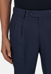 Mann trägt marineblaue Anzughose mit Falten und geknöpftem Bund, Hand locker an der Seite, vor weißem Hintergrund.