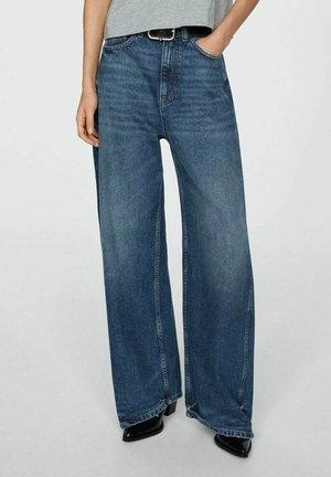 Osoba nosząca niebieskie dżinsy z wysokim stanem i szerokimi nogawkami, czarne botki z ostrym czubkiem, ręce włożone do kieszeni oraz szary top krótszy niż talię.