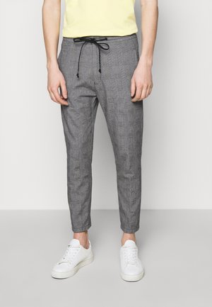Mann trägt graue karierte Slim-Fit-Hose mit schwarzem Kordelzug, weiße Sneakers und ein hellgelbes Hemd, steht vor einem einfarbigen Hintergrund.