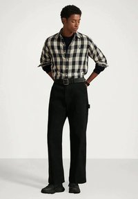 Άνδρας που στέκεται με τα χέρια πίσω από την πλάτη, φορώντας καρό πουκάμισο σε ασπρόμαυρο χρώμα, μαύρο παντελόνι, ζώνη, μαύρα παπούτσια και μαύρο ρολόι.