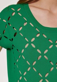 Grünes Strickoberteil mit einem Ausschnitt-Design mit ovalen Formen, einem runden Halsausschnitt und einer glatten Textur, die eine beige Unterlage zeigt.