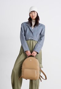 Tanfarbener Leder-Rucksack mit runder Form, Frontreißverschlussfach und verstellbaren Riemen. Getragen von einem Model in einer grauen Jacke und olivgrünen Hosen.