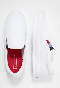 Weiße Slipper-Sneaker aus strukturiertem Stoff, mit einer roten Innensohle und Marineblau/Roten Akzentstreifen an den Seiten, in einer schlanken, abgerundeten Form.