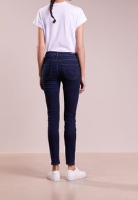 Smala mörka denimjeans med hög midja, kombinerade med en vit t-shirt med kort ärm och vita sneakers, sedd bakifrån.