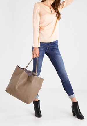 Femme portant un chemisier pêche et un jean bleu, tenant un grand sac fourre-tout en cuir taupe, debout avec une jambe tendue et chaussée de bottines noires.