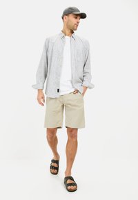 Lys gråstribet langærmet skjorte over en hvid t-shirt, parret med beige shorts og sorte sandaler med to spænder.